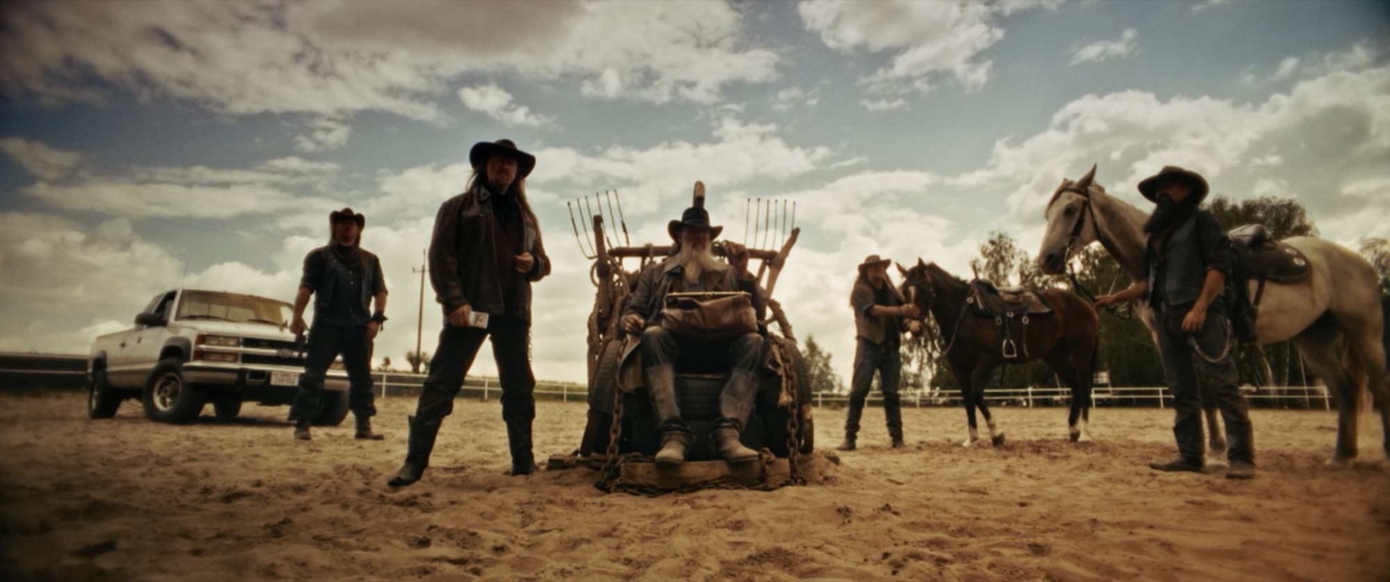 Group portrait in a sandy corral - a bearded elder on a throne of pitchforks and tyres, flanked by cowboys and horses - Amon Amarth